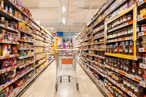 A shopping trolley in a brightly lit supermarket aisle with fully stocked shelves